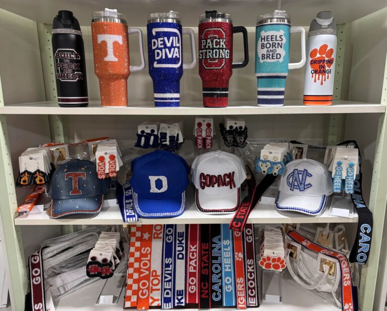Shelf of handmade rhinestone tumblers, denim caps, and earrings for Duke, UNC, NC State, Tennessee, and Clemson at BlingBox
